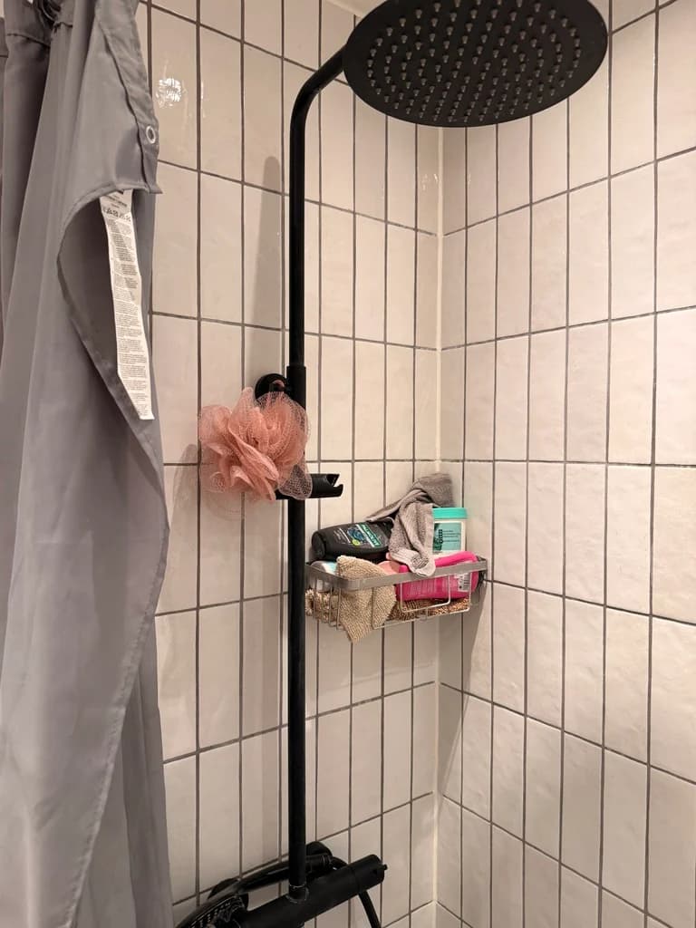 Den gamle bruseniche med små hvide fliser, mørke fuger, sort brusesøjle på væggen og et gråt forhæng.