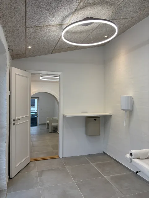 Rum med åben hvid fyldingsdør ind til det næste, en vægmonteret bordplade med dispenser, og cirkulært armatur i akustikloftet.