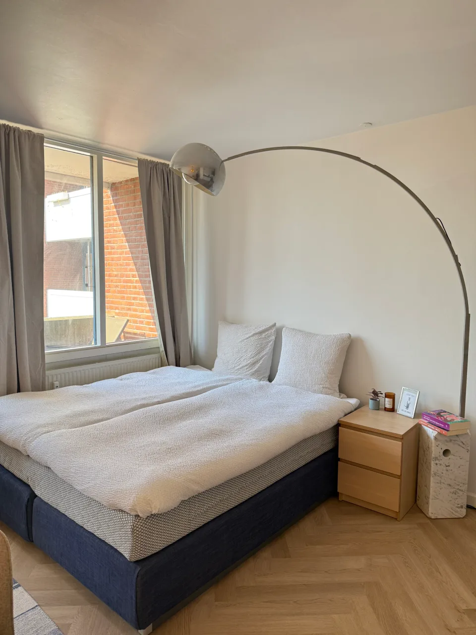 Sovepladsen efter renovering med dobbeltseng, buelampe i stål og natbord i lys eg foran vinduet.